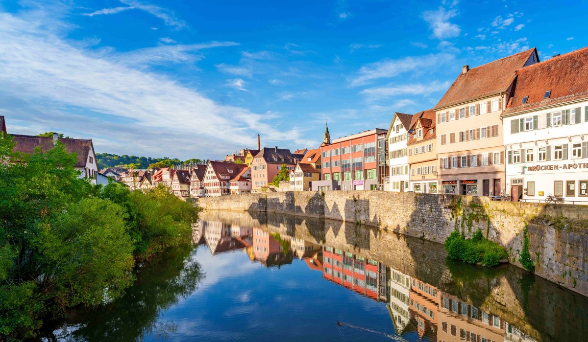Fachwerkhäuser und moderne Gebäude spiegeln sich im Fluss in Schwäbisch Hall. Klare blaue Himmel und grüne Vegetation rahmen die Szene ein., © Michael Kühneisen Fachwerkhäuser und moderne Gebäude spiegeln sich im Fluss in Schwäbisch Hall. Klare blaue Himmel und grüne Vegetation rahmen die Szene ein., © Michael Kühneisen