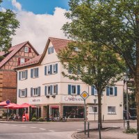 Ein Fachwerkhaus und ein Café mit roten Sonnenschirmen an einer Straßenecke in einer deutschen Stadt, umgeben von Bäumen., © Stadt Welzheim