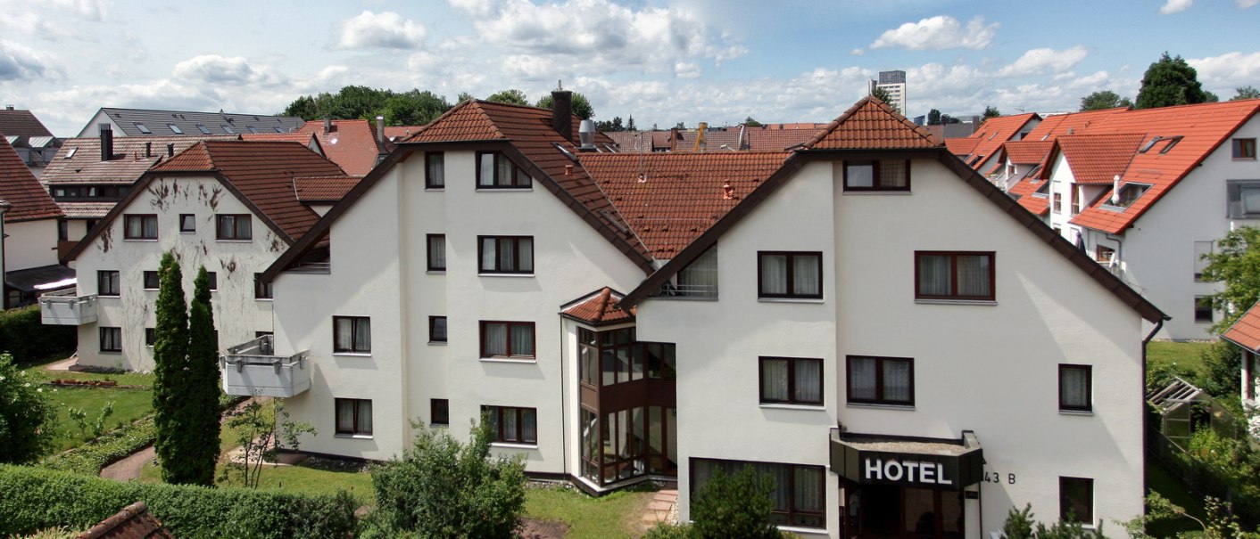 Ein Hotel mit weißer Fassade und roten Dächern, umgeben von Gärten und weiteren Häusern unter einem bewölkten Himmel., © Hotel Flora