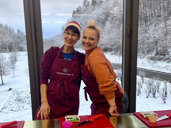 Zwei Frauen in Schürzen stehen in einer Küche. Draußen ist eine schneebedeckte Landschaft zu sehen. Beide lächeln in die Kamera., © Alfred Ritter GmbH & Co. KG | Waldenbuch Zwei Frauen in Schürzen stehen in einer Küche. Draußen ist eine schneebedeckte Landschaft zu sehen. Beide lächeln in die Kamera., © Alfred Ritter GmbH & Co. KG | Waldenbuch