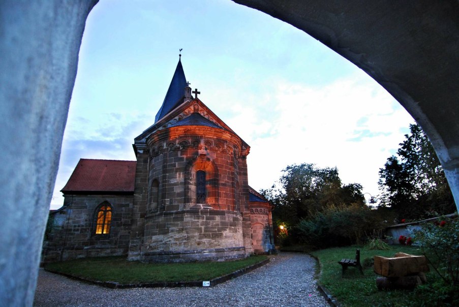 Historische Kirche bei Dämmerung, beleuchtetes Fenster, Kiesweg, Bäume und Bank im Vordergrund.