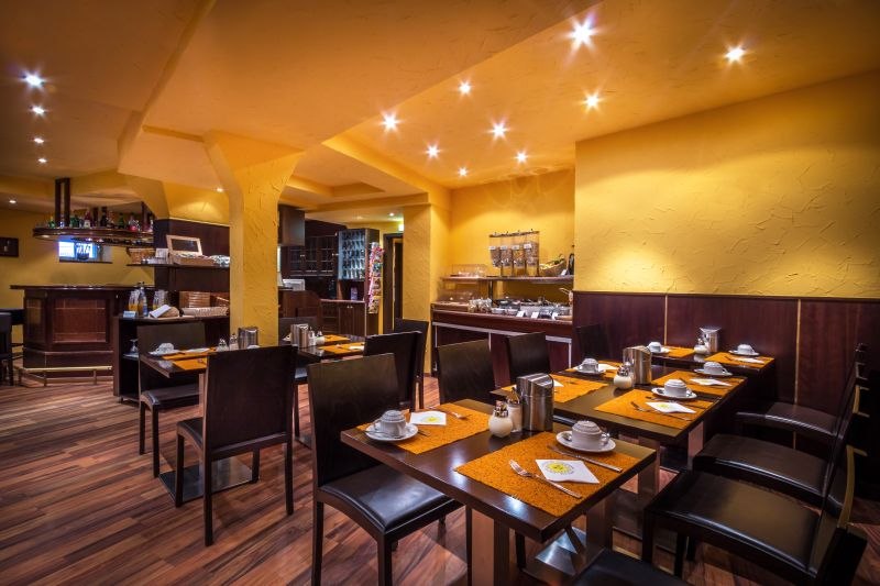 Ein gemütlicher Frühstücksraum mit gedeckten Tischen, warmem Licht und einem Buffet im Hintergrund., © TOMAS