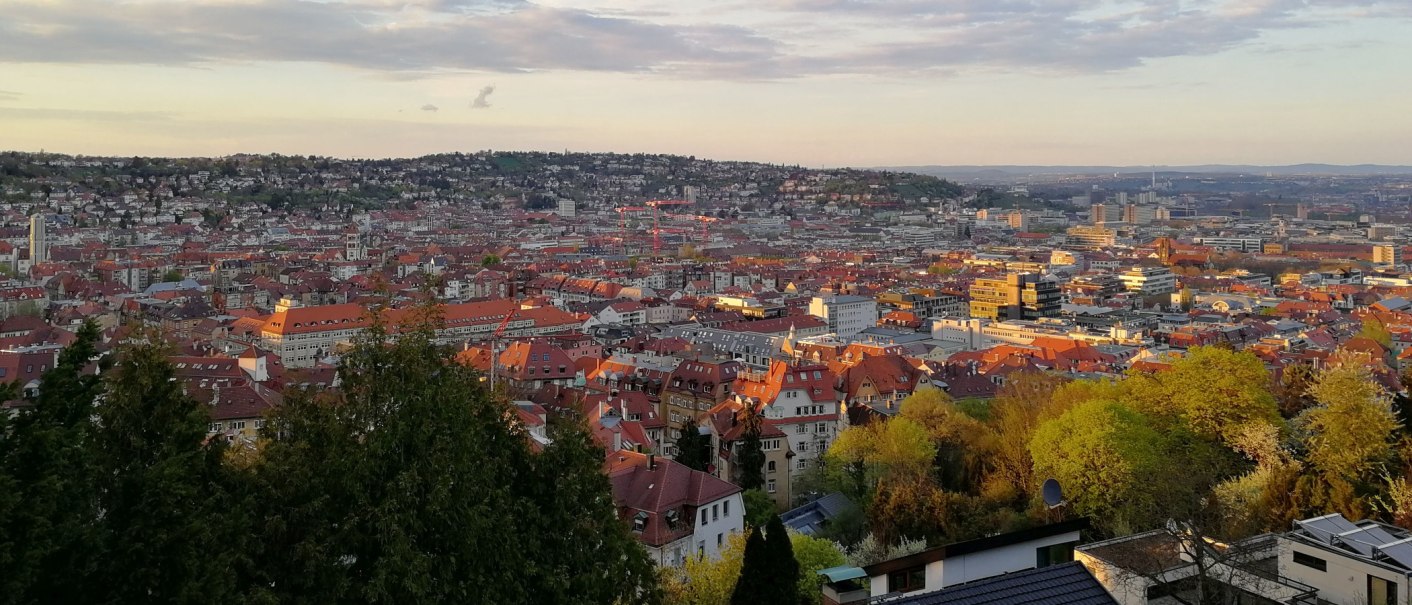 Panoramablick auf Stuttgart bei Sonnenuntergang, mit roten Dächern und grünen Bäumen im Vordergrund. Der Himmel ist leicht bewölkt., © Stuttgart-Marketing GmbH