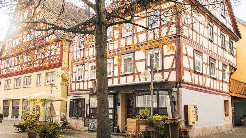 Fachwerkh&auml;user in der Altstadt von K&uuml;nzelsau, umgeben von herbstlichen B&auml;umen. Die Sonne scheint durch die &Auml;ste und beleuchtet die Szene., &copy; Stuttgart-Marketing GmbH, Sarah Schmid