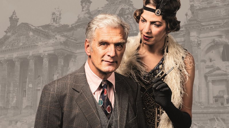 Ein Mann im Anzug und eine Frau im 1920er-Jahre-Kleid stehen vor einem historischen Gebäude. Die Frau trägt eine Feder im Haar.