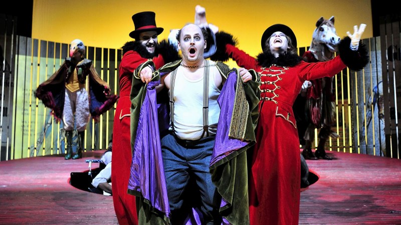 Theaterszene mit Schauspielern in bunten Kost&uuml;men auf einer B&uuml;hne. Ein Mann in rotem Mantel und Zylinder steht im Vordergrund, andere Figuren im Hintergrund., &copy; Patrick Pfeiffer