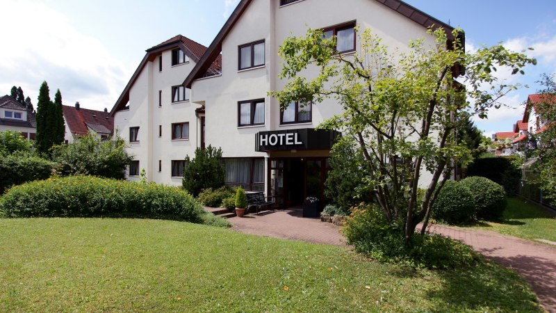 Ein Hotelgebäude mit weißer Fassade und einem Schild "HOTEL" über dem Eingang, umgeben von gepflegtem Garten und Bäumen., © Hotel Flora Ein Hotelgebäude mit weißer Fassade und einem Schild "HOTEL" über dem Eingang, umgeben von gepflegtem Garten und Bäumen., © Hotel Flora