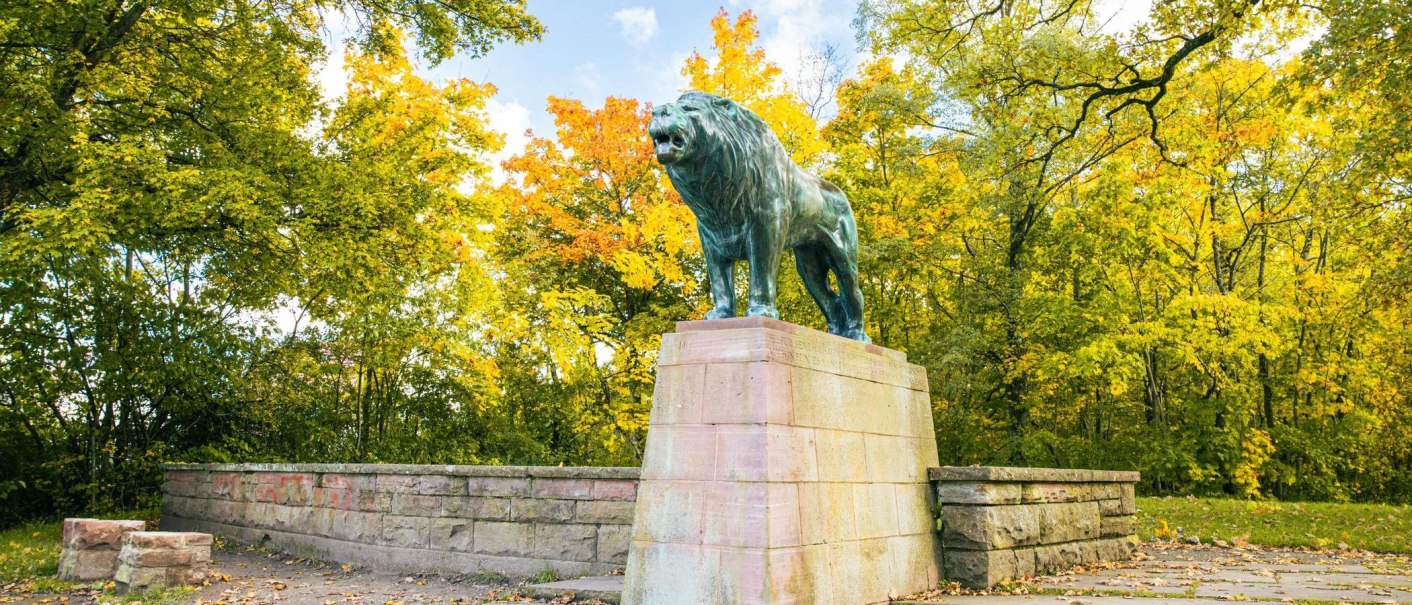 Eine imposante Löwenstatue steht auf einem Sockel, umgeben von herbstlich gefärbten Bäumen in einem Park., © Stuttgart-Marketing GmbH, Sarah Schmid Eine imposante Löwenstatue steht auf einem Sockel, umgeben von herbstlich gefärbten Bäumen in einem Park., © Stuttgart-Marketing GmbH, Sarah Schmid