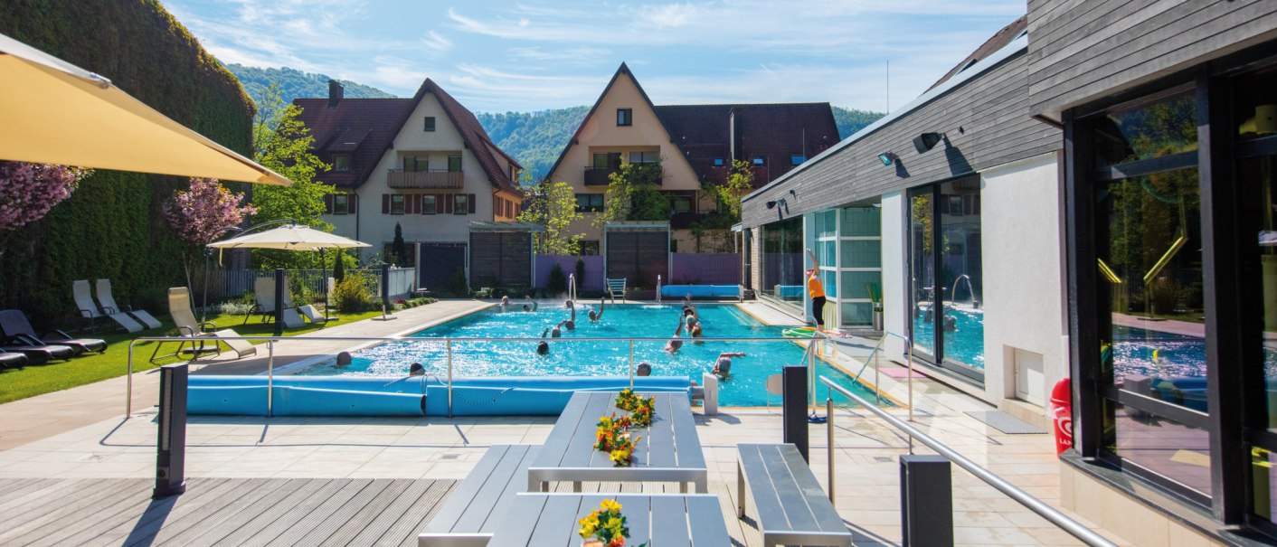 Thermalbad mit Pool, Liegestühlen und Sonnenschirmen. Menschen schwimmen im Wasser, im Hintergrund sind Häuser und grüne Hügel zu sehen., © SMG Achim Mende