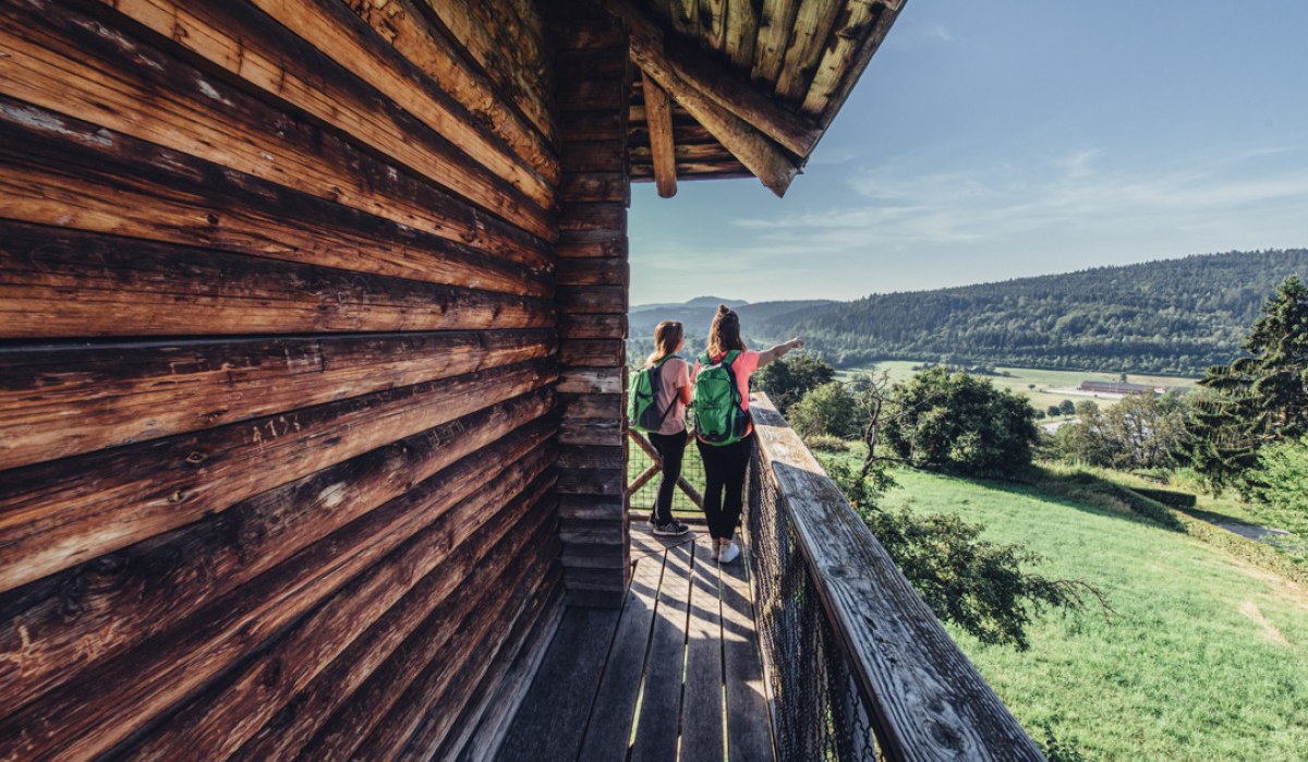 Zwei Personen mit Rucksäcken stehen auf einem Holzbalkon und schauen in die grüne Landschaft. Der Himmel ist klar und blau., © Tourismus Ostalb