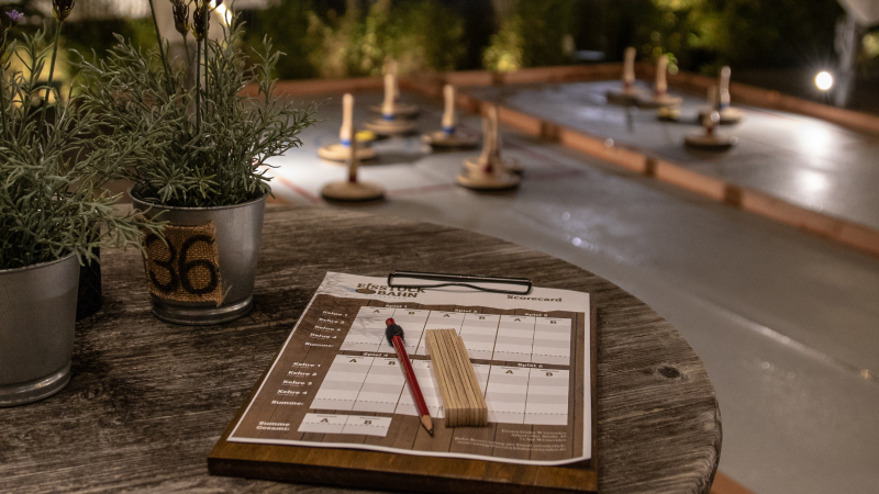 Ein Tisch mit einem Spielplan und Pflanzen im Vordergrund, im Hintergrund eine beleuchtete Eisstockbahn., &copy; 2022 | GaSTra Gastro + Service + Trade GmbH