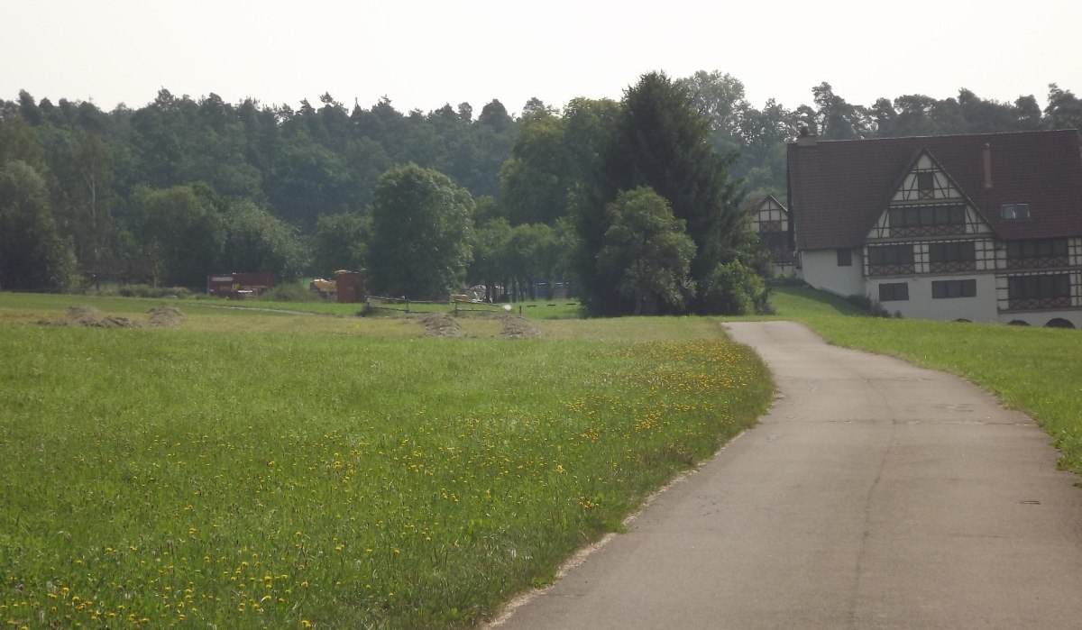Ein asphaltierter Weg führt durch eine grüne Wiese mit gelben Blumen. Im Hintergrund steht ein großes Fachwerkhaus vor einem Wald., © Natur.Nah. Schönbuch & Heckengäu Ein asphaltierter Weg führt durch eine grüne Wiese mit gelben Blumen. Im Hintergrund steht ein großes Fachwerkhaus vor einem Wald., © Natur.Nah. Schönbuch & Heckengäu