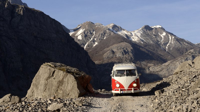 Ein roter VW Bulli auf einem steinigen Bergweg, umgeben von schneebedeckten Bergen unter klarem Himmel., © BLICKFANG Ein roter VW Bulli auf einem steinigen Bergweg, umgeben von schneebedeckten Bergen unter klarem Himmel., © BLICKFANG