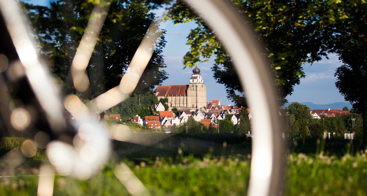 Ein Fahrrad im Vordergrund, durch dessen Speichen eine Kirche in einer Stadt zu sehen ist, umgeben von Bäumen und grüner Landschaft., © Natur.Nah. Schönbuch & Heckengäu Ein Fahrrad im Vordergrund, durch dessen Speichen eine Kirche in einer Stadt zu sehen ist, umgeben von Bäumen und grüner Landschaft., © Natur.Nah. Schönbuch & Heckengäu