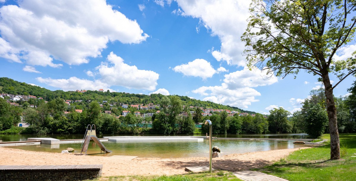 Kocherfreibad Künzelsau: Sandstrand mit Rutsche, umgeben von Bäumen und Hügeln unter blauem Himmel mit Wolken., © Touristikgemeinschaft Hohenlohe, Künzelsau / Stadt Künzelsau Kocherfreibad Künzelsau: Sandstrand mit Rutsche, umgeben von Bäumen und Hügeln unter blauem Himmel mit Wolken., © Touristikgemeinschaft Hohenlohe, Künzelsau / Stadt Künzelsau