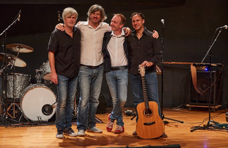 Vier M&auml;nner auf einer B&uuml;hne posieren l&auml;chelnd vor Musikinstrumenten. Einer h&auml;lt eine Gitarre, Schlagzeug im Hintergrund., &copy; Scala Kultur Live gGmbH