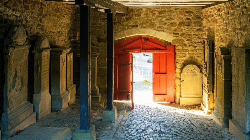Ein historischer Durchgang mit Grabsteinen an den W&auml;nden und einem roten Tor, das nach drau&szlig;en f&uuml;hrt. Sonnenlicht f&auml;llt durch das Tor auf den Boden., &copy; Stuttgart-Marketing GmbH, Sarah Schmid