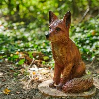 Eine detailreiche Holzskulptur eines Fuchses sitzt auf einem Erlebnispfad im Wald, umgeben von Laub und Pflanzen., © Natur.Nah. Schönbuch & Heckengäu