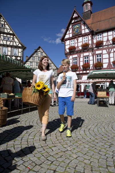 Frau mit Sonnenblumen und Junge mit Eis auf einem Wochenmarkt vor Fachwerkhäusern. Sonniger Tag, Menschen im Hintergrund., © Bad Urach Tourismus Frau mit Sonnenblumen und Junge mit Eis auf einem Wochenmarkt vor Fachwerkhäusern. Sonniger Tag, Menschen im Hintergrund., © Bad Urach Tourismus