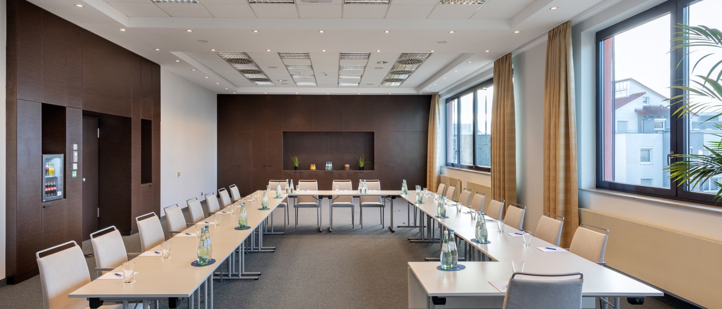 Moderner Konferenzraum mit U-förmiger Tischanordnung, Stühlen und Wasserflaschen. Große Fenster lassen viel Licht herein, braune Vorhänge und Pflanzen dekorieren den Raum., © NH Hotel Stuttgart Airport Moderner Konferenzraum mit U-förmiger Tischanordnung, Stühlen und Wasserflaschen. Große Fenster lassen viel Licht herein, braune Vorhänge und Pflanzen dekorieren den Raum., © NH Hotel Stuttgart Airport
