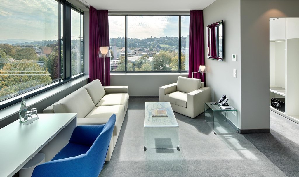 Moderne Suite mit großen Fenstern, Sofa, Sessel, Schreibtisch und Stadtblick. Helle Farben und stilvolle Einrichtung dominieren den Raum., © Hotel am Remspark Moderne Suite mit großen Fenstern, Sofa, Sessel, Schreibtisch und Stadtblick. Helle Farben und stilvolle Einrichtung dominieren den Raum., © Hotel am Remspark