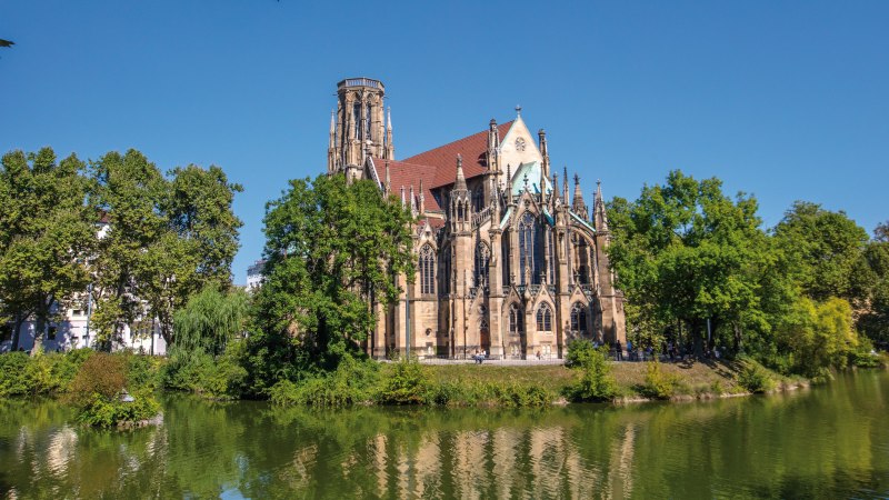 Die Johanneskirche am Feuersee in Stuttgart, umgeben von Bäumen und reflektiert im Wasser, bei klarem, sonnigem Wetter., © SMG Achim Mende Die Johanneskirche am Feuersee in Stuttgart, umgeben von Bäumen und reflektiert im Wasser, bei klarem, sonnigem Wetter., © SMG Achim Mende