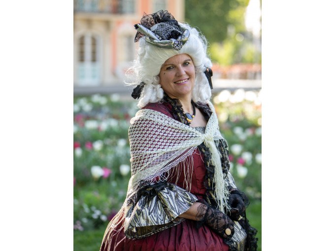 Eine Frau in barockem Kost&uuml;m mit wei&szlig;er Per&uuml;cke und rotem Kleid steht l&auml;chelnd im Freien vor einem Blumenbeet., &copy; Touristik und Marketing GmbH
