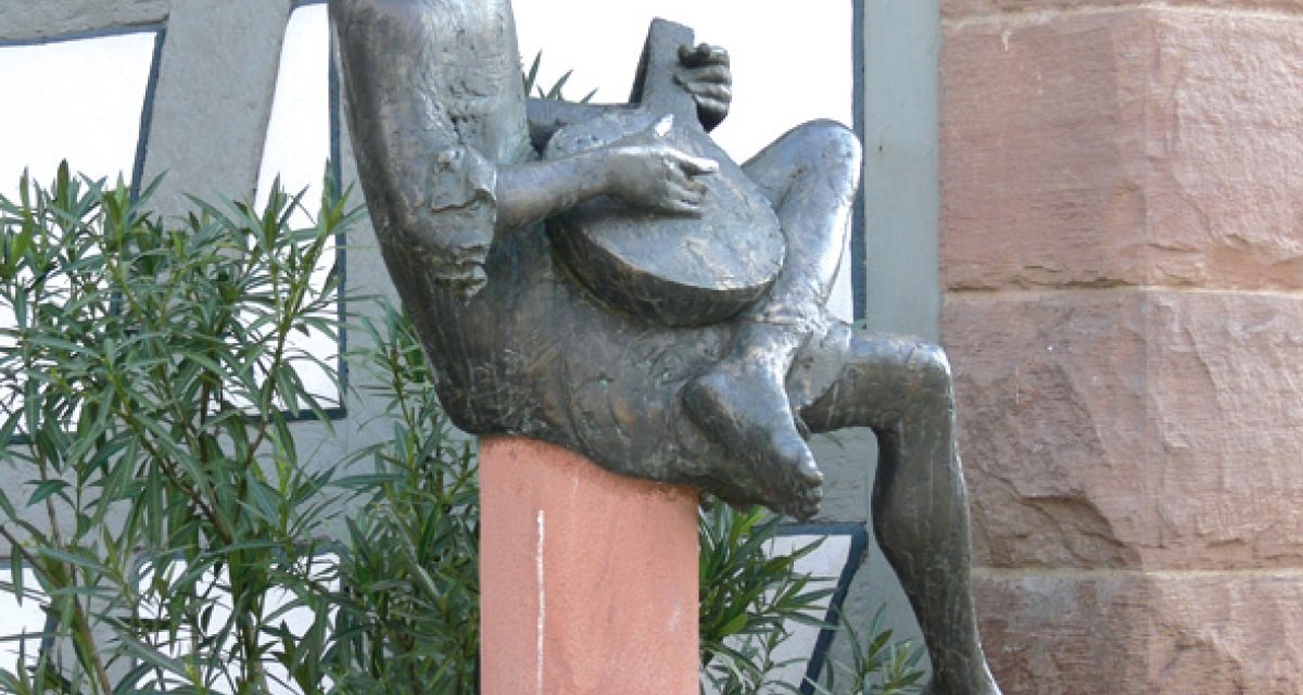 Bronzestatue eines Minnesängers mit Laute, sitzend auf einem Sockel vor einem Fachwerkgebäude. Im Hintergrund sind Pflanzen und eine Steinmauer zu sehen., © Land der 1000 Hügel - Kraichgau-Stromberg Bronzestatue eines Minnesängers mit Laute, sitzend auf einem Sockel vor einem Fachwerkgebäude. Im Hintergrund sind Pflanzen und eine Steinmauer zu sehen., © Land der 1000 Hügel - Kraichgau-Stromberg