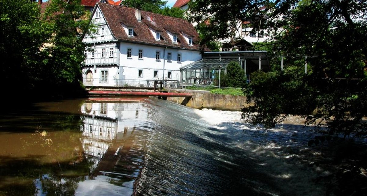 Historische Mühle mit Fachwerk und rotem Dach an einem Fluss mit Wehranlage, umgeben von Bäumen und blauem Himmel., © Waiblingen - Stuttgart-Marketing GmbH Historische Mühle mit Fachwerk und rotem Dach an einem Fluss mit Wehranlage, umgeben von Bäumen und blauem Himmel., © Waiblingen - Stuttgart-Marketing GmbH