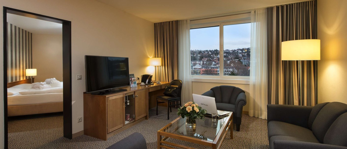 Elegante Junior Suite mit Wohnzimmer, Sofa, Tisch, Fernseher und großem Fenster mit Stadtblick. Schlafzimmer im Hintergrund sichtbar., © Maritim Hotelgesellschaft mbH