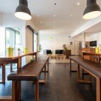 Moderne Lobby mit Holztischen und Bänken, großen Lampen und einem Sitzbereich im Hintergrund. Helle, einladende Atmosphäre., © a2 HOTELS