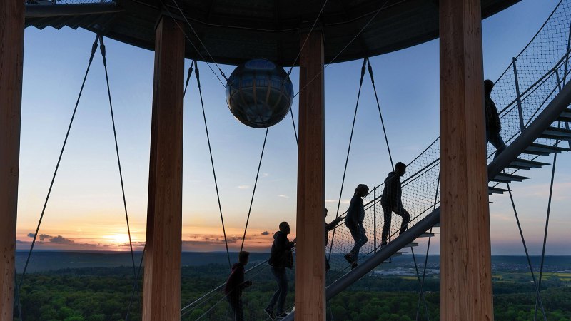 Menschen steigen eine Wendeltreppe im Schönbuchturm hinauf, umgeben von Holzsäulen. Im Hintergrund ist ein Sonnenuntergang über einer weiten Landschaft zu sehen., © Stuttgart-Marketing GmbH, Martina Denker