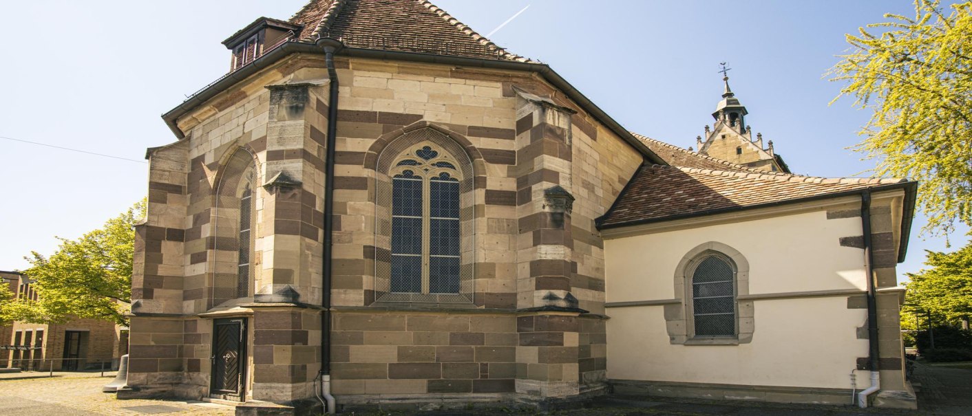Die Lutherkirche in Fellbach mit gotischen Fenstern und gemusterten Steinwänden, umgeben von Bäumen unter klarem Himmel., © SMG, Sarah Schmid