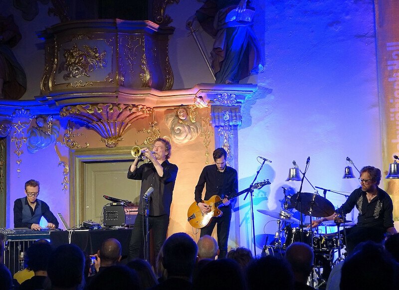 Das Nils Petter Molvær Trio performt auf einer Bühne mit barocker Dekoration. Die Musiker spielen Trompete, Gitarre und Schlagzeug vor einem interessierten Publikum., © Jazzclub Schwäbisch Hall e.V. Das Nils Petter Molvær Trio performt auf einer Bühne mit barocker Dekoration. Die Musiker spielen Trompete, Gitarre und Schlagzeug vor einem interessierten Publikum., © Jazzclub Schwäbisch Hall e.V.
