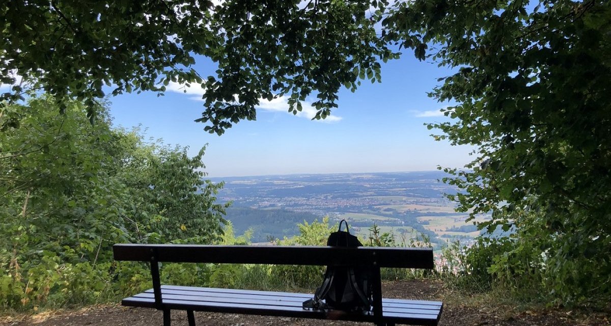 Eine Bank mit Rucksack steht unter Bäumen, bietet einen weiten Blick auf die Landschaft. Der Himmel ist blau mit wenigen Wolken., © Touristik und Marketing GmbH Eine Bank mit Rucksack steht unter Bäumen, bietet einen weiten Blick auf die Landschaft. Der Himmel ist blau mit wenigen Wolken., © Touristik und Marketing GmbH
