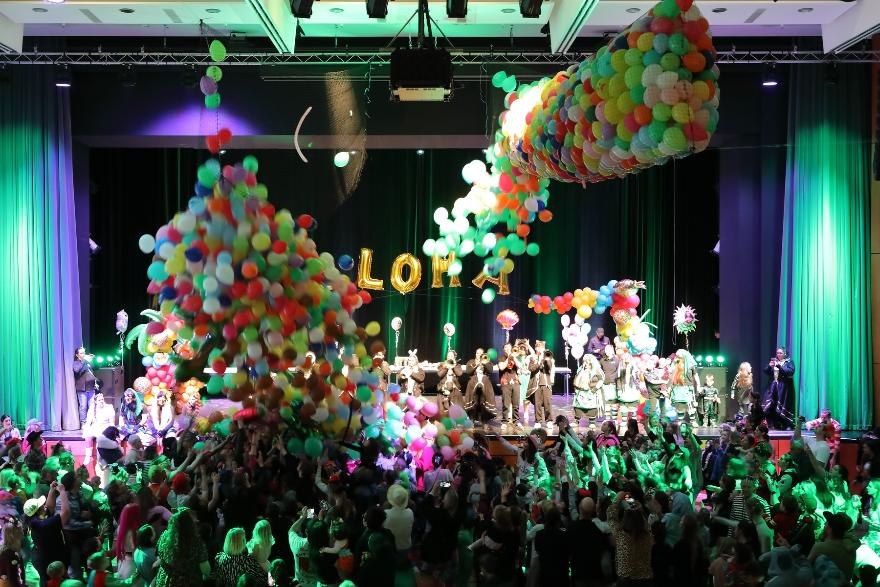 Bunte Ballons fallen von der Decke auf eine fröhliche Menschenmenge bei einer Kinderfaschingsveranstaltung. Bühne mit verkleideten Personen im Hintergrund., © © Veranstalter
