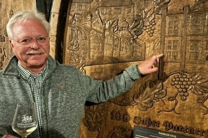 Ein Mann zeigt auf ein kunstvoll geschnitztes Weinfass in einem Weinkeller. Er hält ein Weinglas und lächelt in die Kamera., © Weingenuss Stuttgart