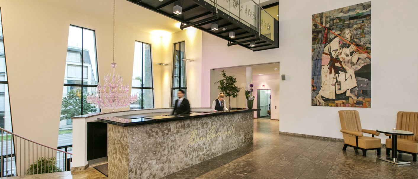 Moderne Hotelrezeption mit Marmortresen, großem Kronleuchter, Kunstwerk an der Wand und zwei Angestellten. Helle, stilvolle Einrichtung., © ARCOTEL Hotels