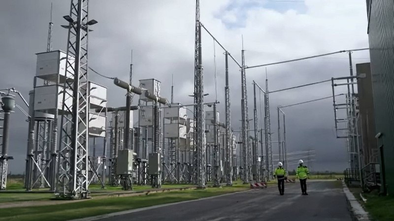 Zwei Personen in gelben Westen gehen durch ein Umspannwerk mit zahlreichen Strommasten und grauem Himmel., © Kulturhaus Schwanen