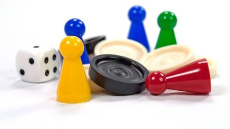 Bunte Spielfiguren und ein Würfel liegen verstreut auf weißem Hintergrund. Die Figuren sind in Gelb, Blau, Grün und Rot., © Stadtbibliothek Ludwigsburg