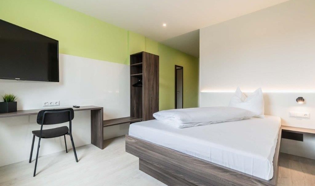 Modernes Einzelzimmer mit Bett, Schreibtisch, Stuhl und Fernseher. Die Wände sind in Grün und Weiß gehalten, das Mobiliar ist aus dunklem Holz., © a2 HOTELS am Quadrium