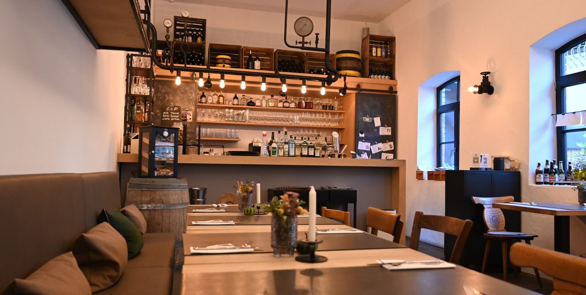 Gem&uuml;tliches Restaurant mit Bar, Holztischen und -st&uuml;hlen, dekorativen Elementen und warmem Licht. Die Bar ist gut best&uuml;ckt mit Flaschen und Gl&auml;sern.