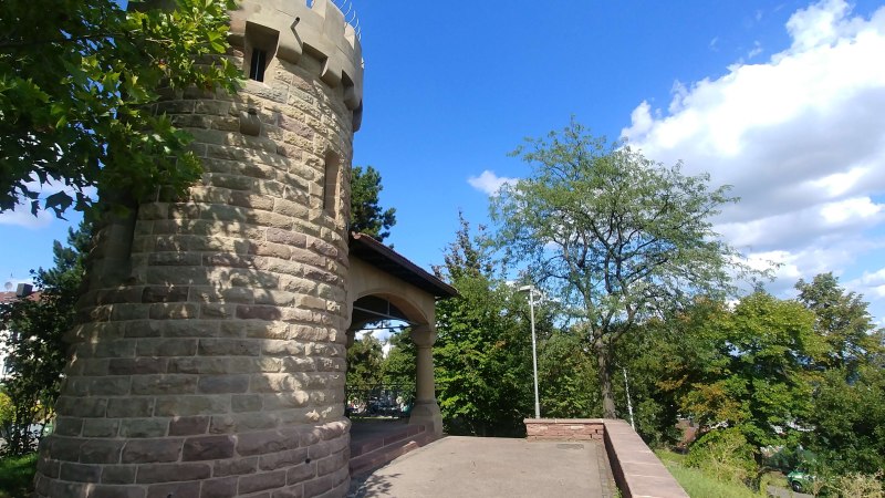 Runder Steinturm mit Zinnen, umgeben von Bäumen und blauem Himmel. Der Turm steht auf einer gepflasterten Fläche., © SMG