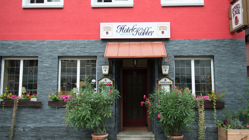 Das Hotel Köhler hat eine rote Fassade mit grauen Steinelementen. Vor dem Eingang stehen Pflanzen und bunte Blumen schmücken die Fenster., © Hotel Köhler Das Hotel Köhler hat eine rote Fassade mit grauen Steinelementen. Vor dem Eingang stehen Pflanzen und bunte Blumen schmücken die Fenster., © Hotel Köhler