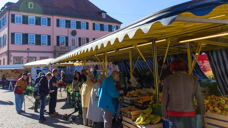 Menschen kaufen auf einem Markt in Murrhardt ein. Gelbe Marktstände bieten Obst und Gemüse an. Ein großes, rosa Gebäude im Hintergrund., © Stuttgart-Marketing GmbH Menschen kaufen auf einem Markt in Murrhardt ein. Gelbe Marktstände bieten Obst und Gemüse an. Ein großes, rosa Gebäude im Hintergrund., © Stuttgart-Marketing GmbH