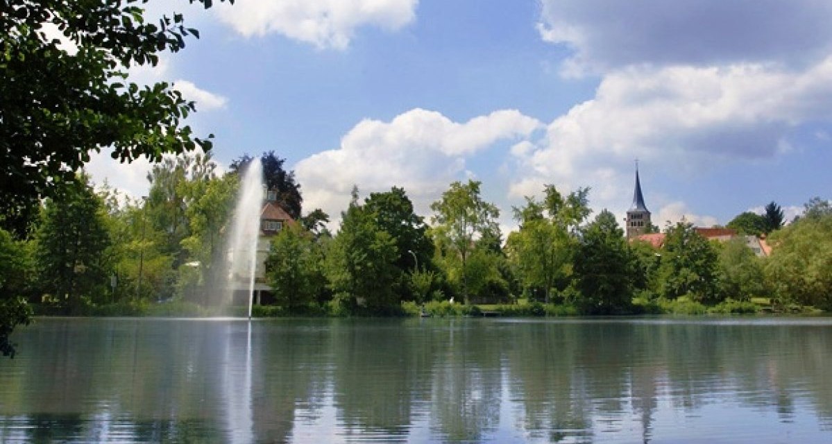 Ein idyllischer See mit einem Springbrunnen, umgeben von Bäumen. Im Hintergrund ist ein Kirchturm zu sehen, der in den blauen Himmel ragt., © Sindelfingen - Stuttgart-Marketing GmbH Ein idyllischer See mit einem Springbrunnen, umgeben von Bäumen. Im Hintergrund ist ein Kirchturm zu sehen, der in den blauen Himmel ragt., © Sindelfingen - Stuttgart-Marketing GmbH