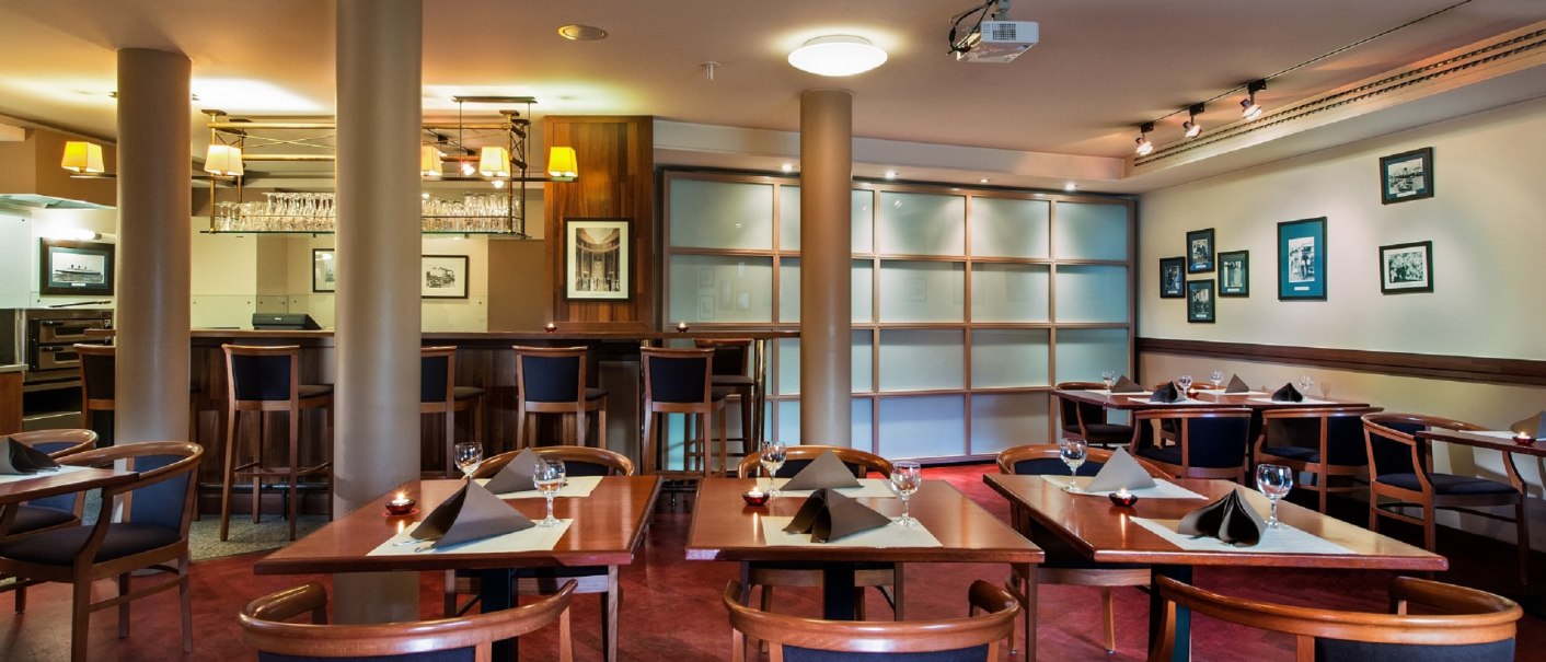 Elegantes Restaurant mit Holzmöbeln, gedeckten Tischen und Barbereich. Dezente Beleuchtung und dekorative Bilder an den Wänden., © TOMAS