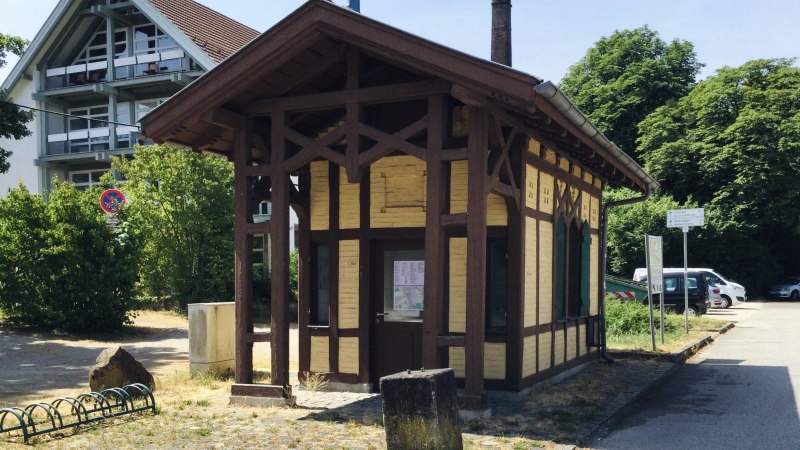 Kleines Fachwerkgebäude am Neckartalradweg, umgeben von Bäumen und einem modernen Gebäude. Ein Infopunkt mit Schildern und Fahrradständer., © J. Tubolzer