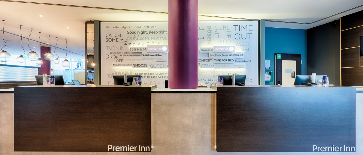 Moderne Hotelrezeption mit zwei Schaltern, lila Säule und Premier Inn Logo. Im Hintergrund eine Wand mit Schlafmotiven., © Premier Inn Germany