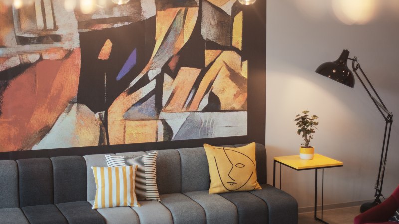 Moderne Lobby mit grauem Sofa, bunten Kissen, abstraktem Wandbild, gelbem Beistelltisch, Pflanze und Stehlampe., &copy; the niu Form - Novum Hospitality